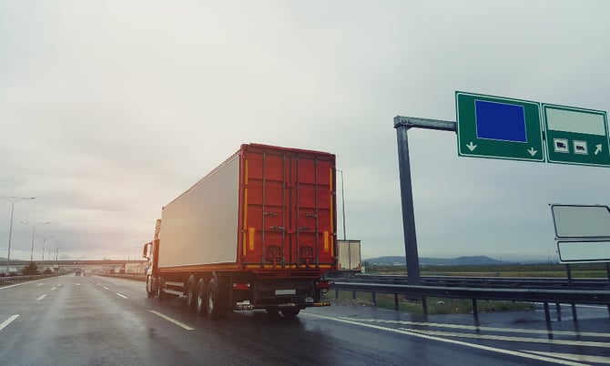 A large red truck drives along a motorway towards the horizon.