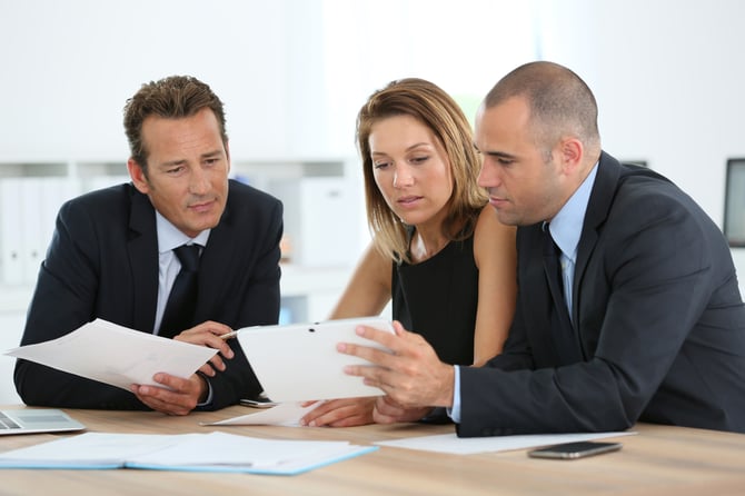 Three people in suits examine paperwork at a desk.
