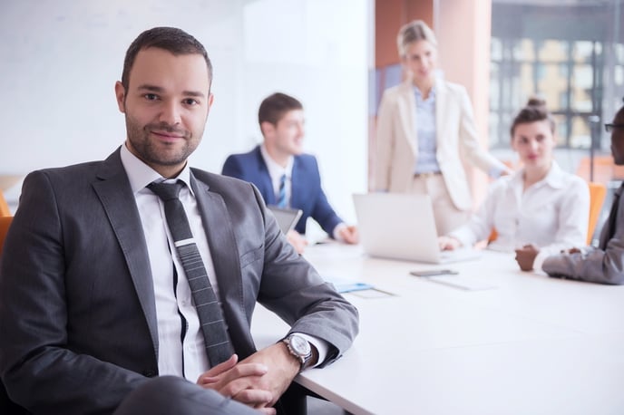 A businessman sits at a long desk and smiles at the camera. Behind other people look at a laptop.